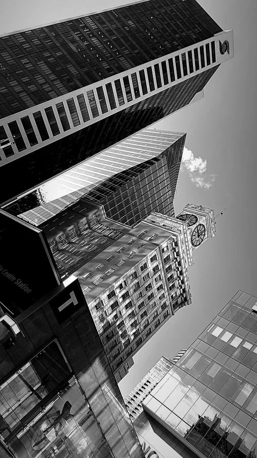 Looking up at converging skyscrapers and a heritage clock tower in black and white
